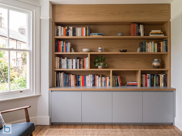A custom built-in contemporary alcove bookcase featuring seamless light grey handless base cupboards beneath an expansive, floor-to-ceiling natural oak display shelving unit filled with books.