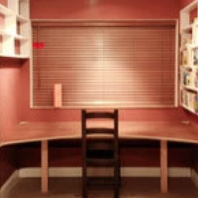 Fitted home office desk built into a red alcove with open display shelving above, a roller blind, and a wooden chair positioned in front of the desk.
