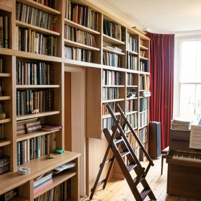 Fitted oak bookcases with a built in library ladder for reaching upper shelves, beside a window with red curtains.