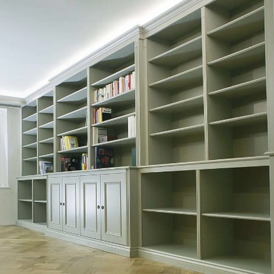 A long, floor to ceiling fitted bookcase painted sage green, with open shelving for books above closed cupboards along the full length of the wall.