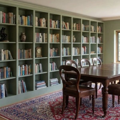 Floor to ceiling fitted bookcases painted in sage green, lining a dining room wall behind a wooden table and chairs.