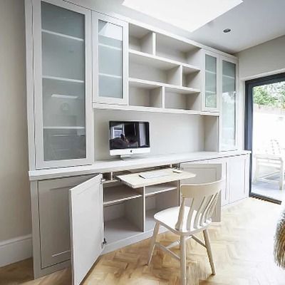 Grey fitted home office cabinets with glass fronted display doors above open cupboards, a pull out desk with computer, and a wooden chair beneath a skylight.