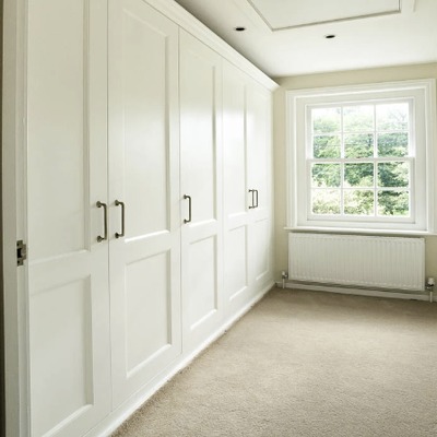 A long run of fitted wardrobes with cream shaker doors and chrome bar handles, fitted along a hallway landing beside a window.