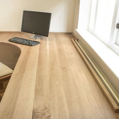 Fitted oak desk with a computer monitor and keyboard positioned in a corner beside a large window, showing the natural wood grain of this full length plank desk