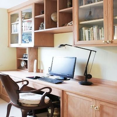 Cherry wood fitted desk with computer monitor and lamp, beneath glass fronted wall cabinets holding books and ornaments, with a leather armchair positioned at the desk.