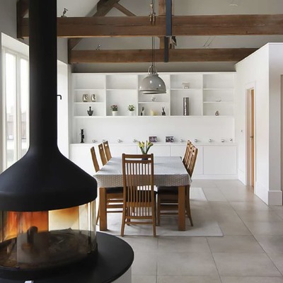 An open plan kitchen and dining area with white fitted shelving along the back wall, exposed oak beams, and a central black wood burning stove.