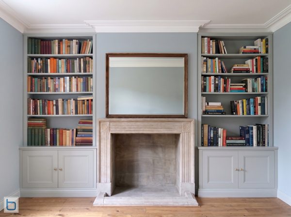 A pair of custom-built grey alcove units featuring traditional shaker base cupboards and open upper bookshelves, symmetrically flanking a large stone fireplace and mirror.