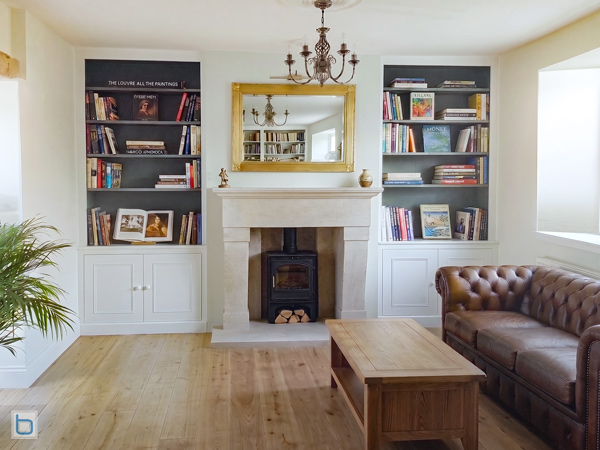 A pair of custom-built white alcove units flanking a stone fireplace mantle, featuring traditional shaker base cupboards and open upper bookshelves with contrasting dark painted backing panels.