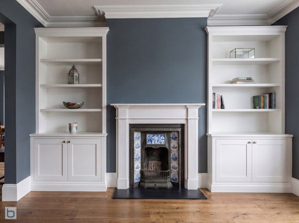 A pair of custom-built white alcove units with traditional shaker base cupboards and open upper shelving, flanking a classic period fireplace mantel in a room with deep grey walls and oak flooring.
