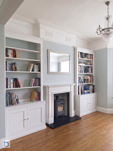A pair of bespoke white alcove units featuring traditional shaker base cupboards and open upper bookshelves, flanking a white mantelpiece with a log burner stove against a duck egg blue wall.