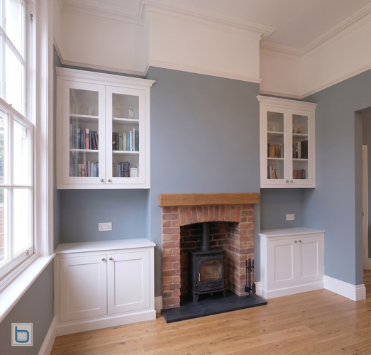 A pair of custom-built grey alcove units featuring traditional shaker base cupboards and open upper bookshelves, symmetrically flanking a large stone fireplace and mirror.