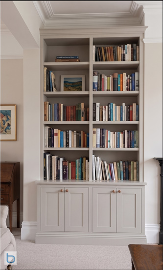 A custom-built transitional alcove bookcase painted in a soft greige finish, featuring a four-door shaker base cabinet with small knob handles and an open upper bookcase filled with books.