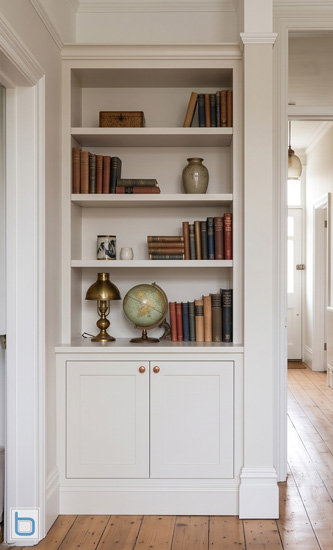 A custom-built transitional alcove unit painted in a soft off-white finish, featuring a two-door shaker base cabinet with brass knobs and upper display shelves styled with books, a globe, and a vintage lamp.