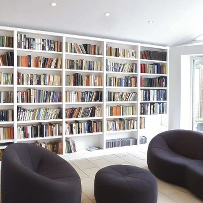 White fitted bookcases filled with books lining a bright garden room, furnished with brown curved sofas and footstools.