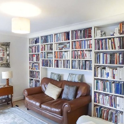 White fitted bookcases filled with books lining a living room wall, furnished with a brown leather sofa and fireplace.