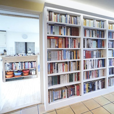 White fitted bookcases filled with books lining a hallway wall, leading through to a kitchen with tiled flooring.