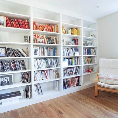White fitted bookcases with open shelving holding a large book collection, positioned beside a cream sofa on wooden flooring.