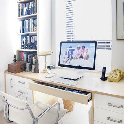 White fitted desk with pull out drawer built into a bay window, bookshelves above holding books and a globe, with a computer monitor and desk lamp.