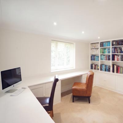 White fitted bookshelves and cupboards beside a long desk built under a window, with a dark leather office chair and a tan armchair in the room.