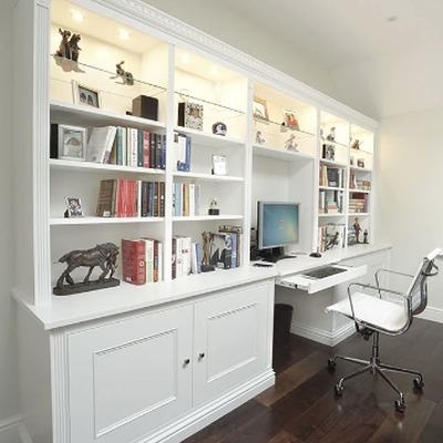 White fitted home office with floor to ceiling display shelving, storage cupboards below, a central desk with computer, and a chair on dark oak flooring.