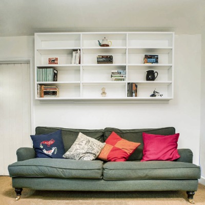 White fitted wall mounted shelving displaying books and ornaments above a green sofa with colourful scatter cushions.