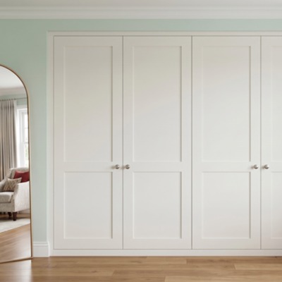White fitted wardrobes with round knob handles, fitted along a wall in a bedroom with sage green walls and wood flooring.