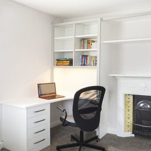 White painted fitted home office with desk, open shelving and base drawers beside a period fireplace
