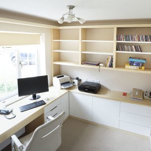 Modern white fitted home office with L-shaped desk, overhead shelving and base cupboards, natural light from window