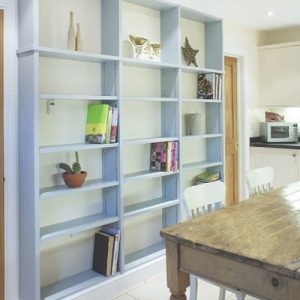 Pale blue fitted shelving displaying books and ornaments in a kitchen diner, positioned beside a farmhouse style table.