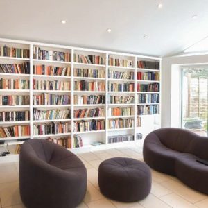 White fitted bookcases filled with books lining a bright garden room, furnished with curved dark sofas and footstools.