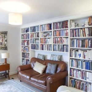 White fitted bookcases filled with books lining a living room wall, furnished with a brown leather sofa and fireplace.